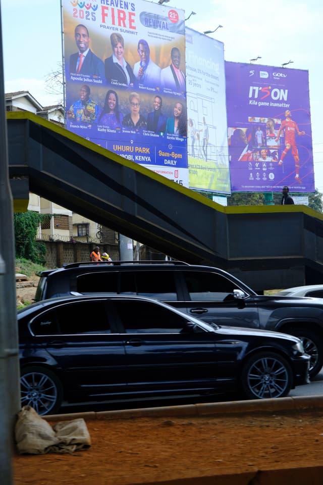 A street scene with parked cars in the foreground and two large billboards in the background, one advertising an event and the other promoting a product. An overpass or pedestrian bridge is visible above the cars