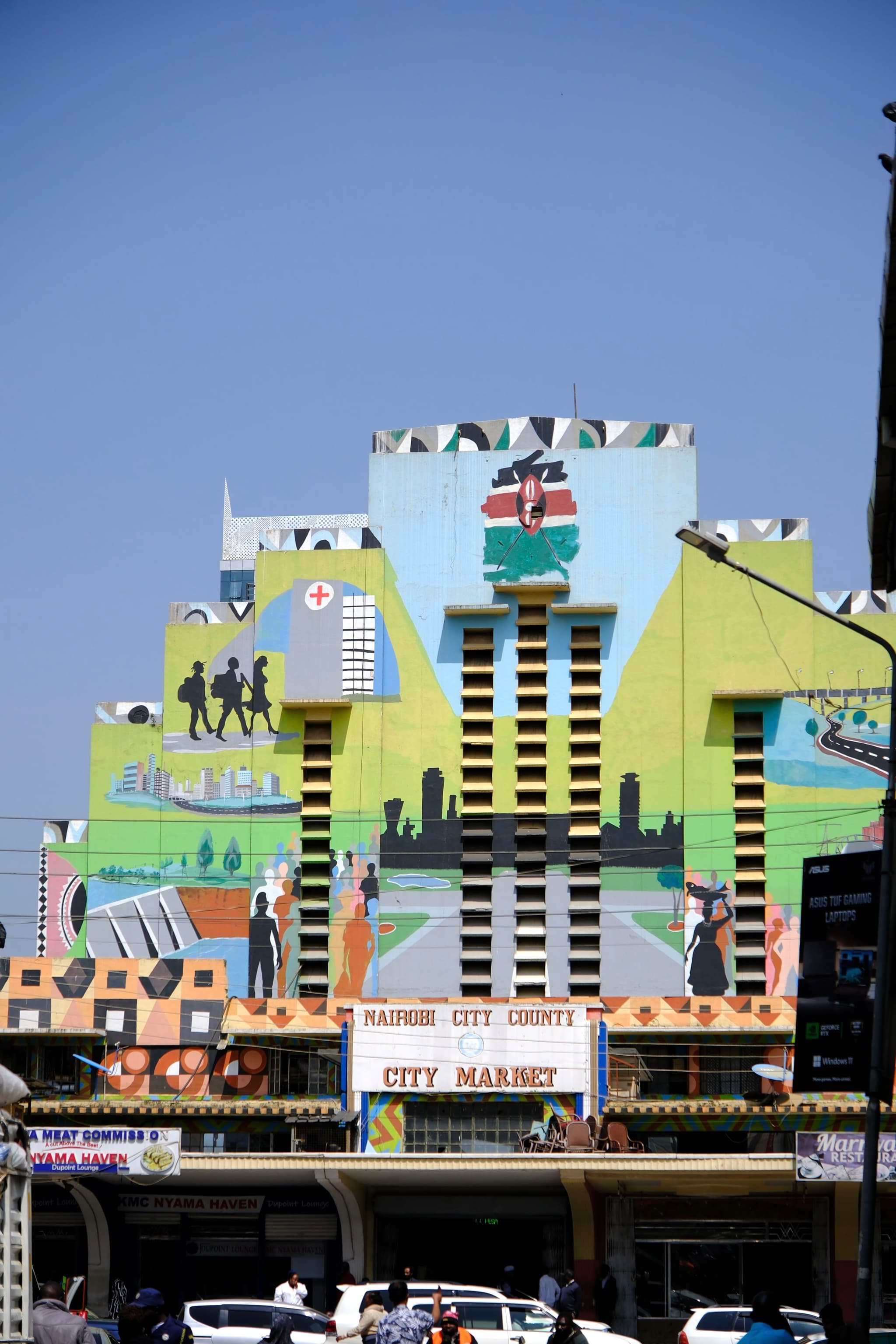 A colorful building facade with murals depicting various scenes and the Kenyan flag, located in a busy urban area with cars and people