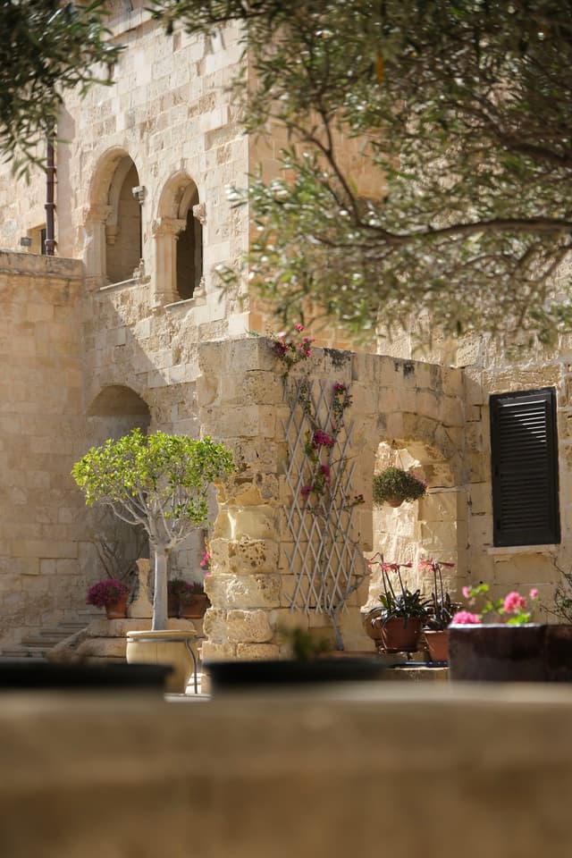 A stone building with arched windows, surrounded by potted plants and trees, creating a serene courtyard atmosphere