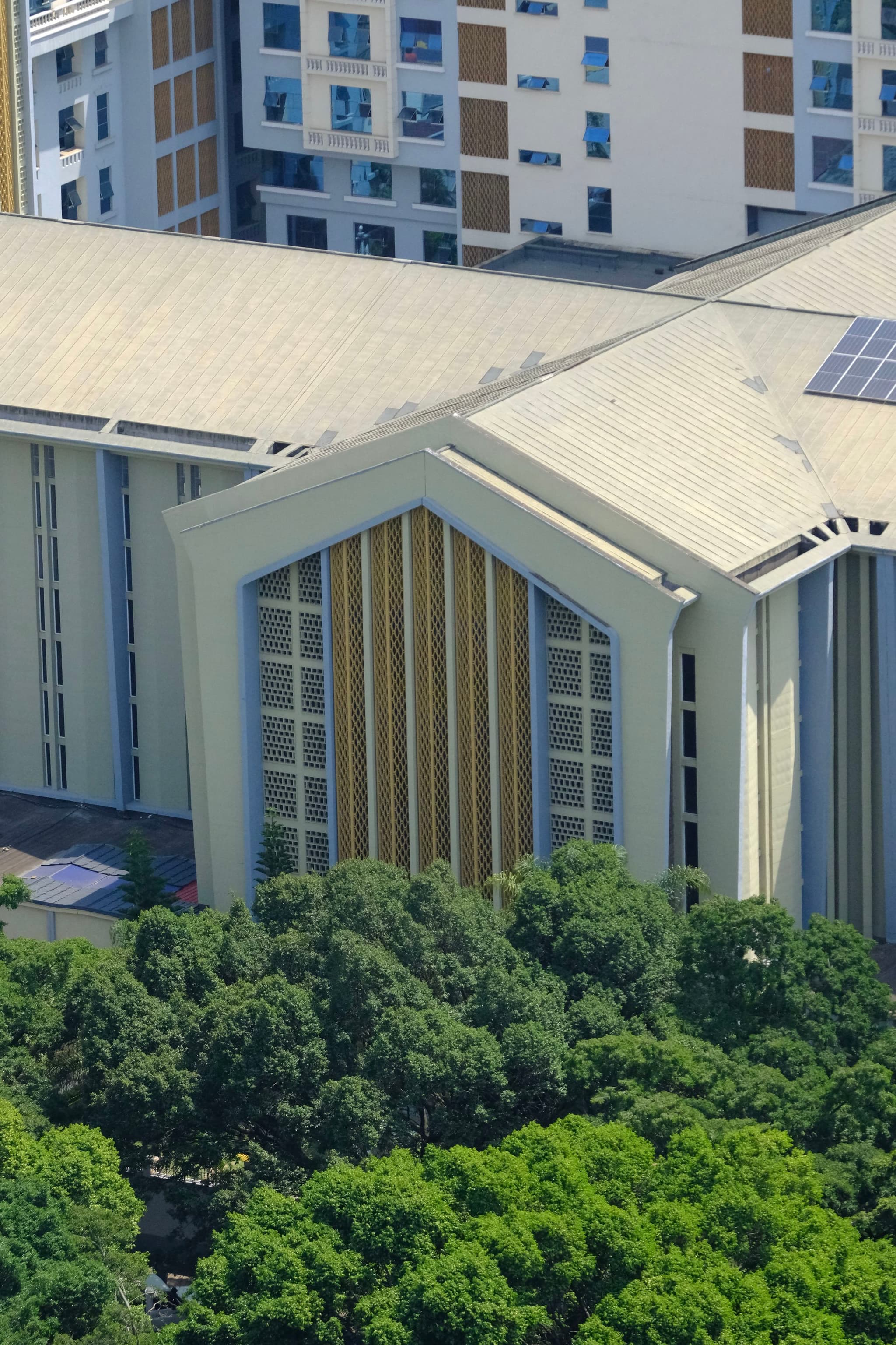 A modern building with a large, angular roof and vertical window panels, surrounded by lush green trees