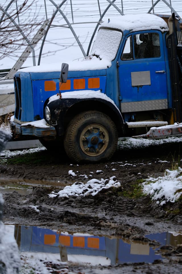 A parked blue vintage truck dusted with snow beside muddy ground and a reflective puddle near metal greenhouse frames