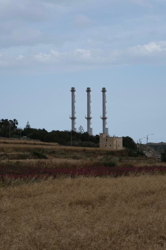Three tall industrial chimneys rise above a landscape of dry grass and fields, with a small building at their base and a partly cloudy sky overhead