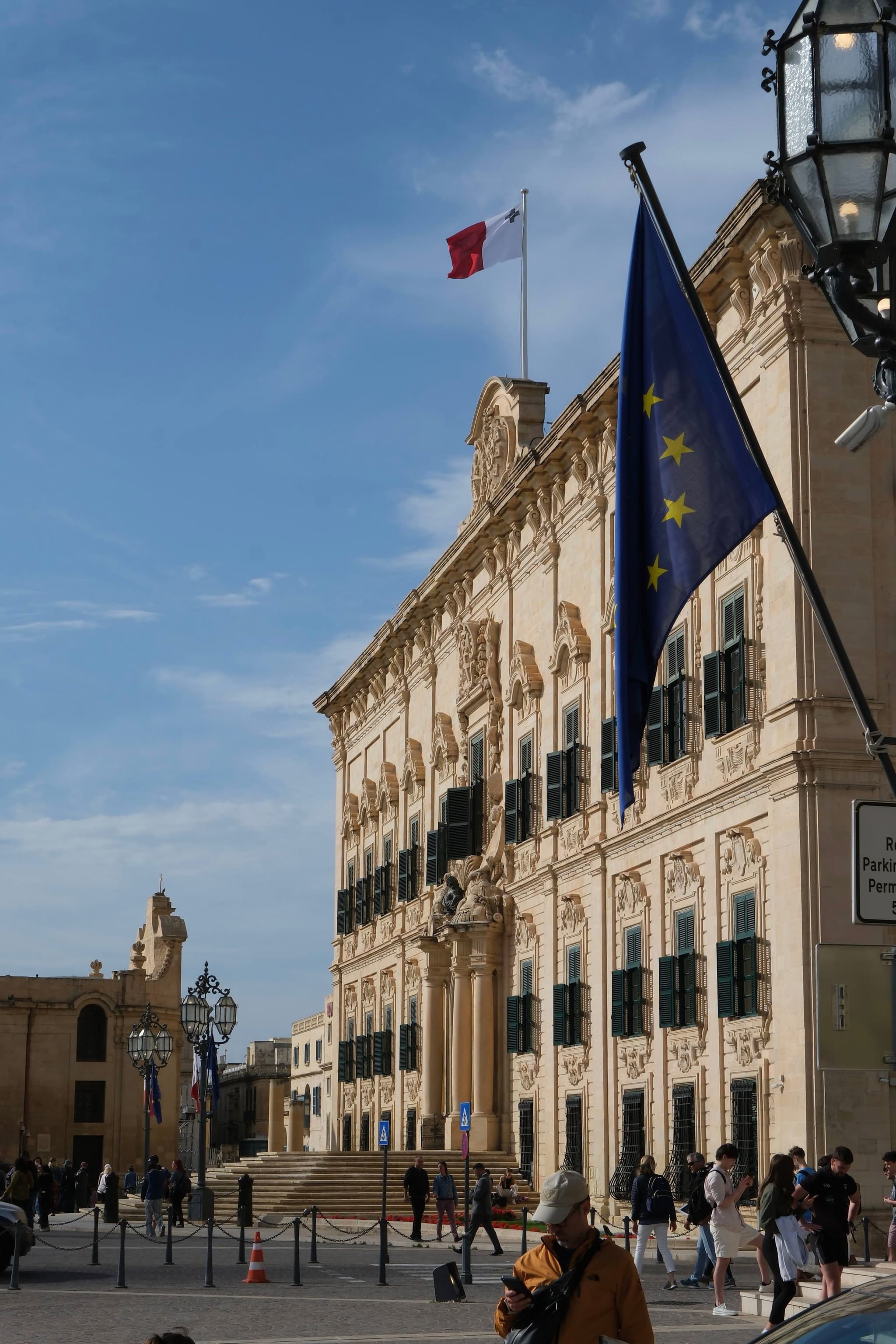 A historic building with ornate architecture, featuring a Maltese flag on top and an EU flag in the foreground, under a clear blue sky. People are walking nearby
