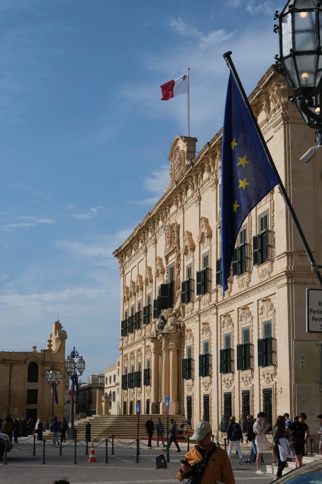 A historic building with ornate architecture, featuring a Maltese flag on top and an EU flag in the foreground, under a clear blue sky. People are walking nearby