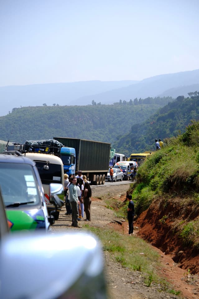 A traffic jam on a rural road with vehicles lined up and people standing outside. The background features rolling hills and a clear sky