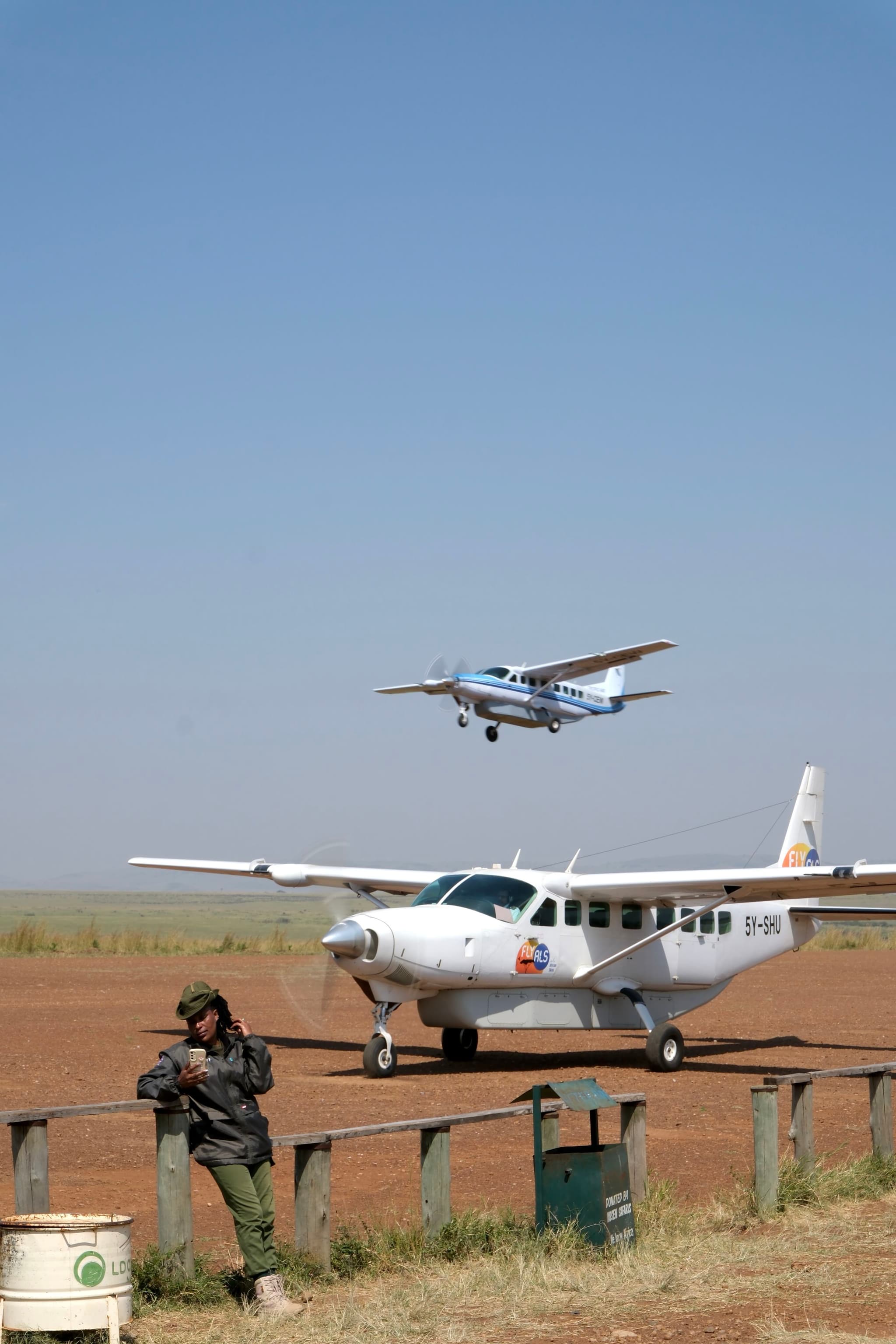 A small aircraft is parked on a dirt airstrip with another plane flying overhead. A person stands nearby, leaning on a fence, under a clear blue sky
