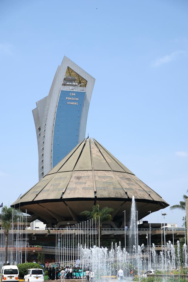 A modern, angular skyscraper stands behind a large, dome-shaped building with a fountain in the foreground