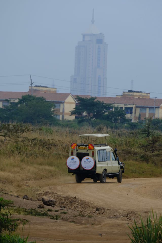 A vehicle with two large drums on the back is driving on a dirt road, with a tall building and smaller structures in the background