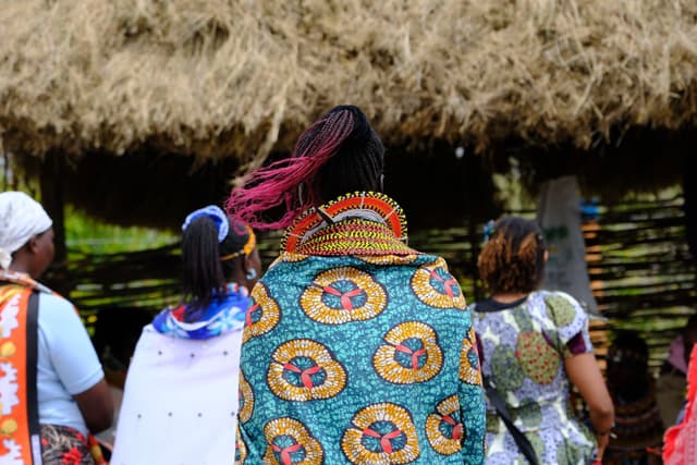 A group of people wearing colorful traditional clothing and accessories, standing in front of a thatched structure