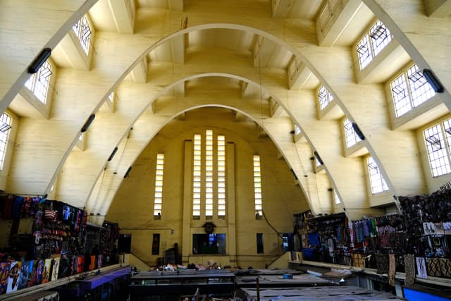 An interior view of a large, arched market hall with high ceilings, featuring rows of windows and various stalls lining the sides