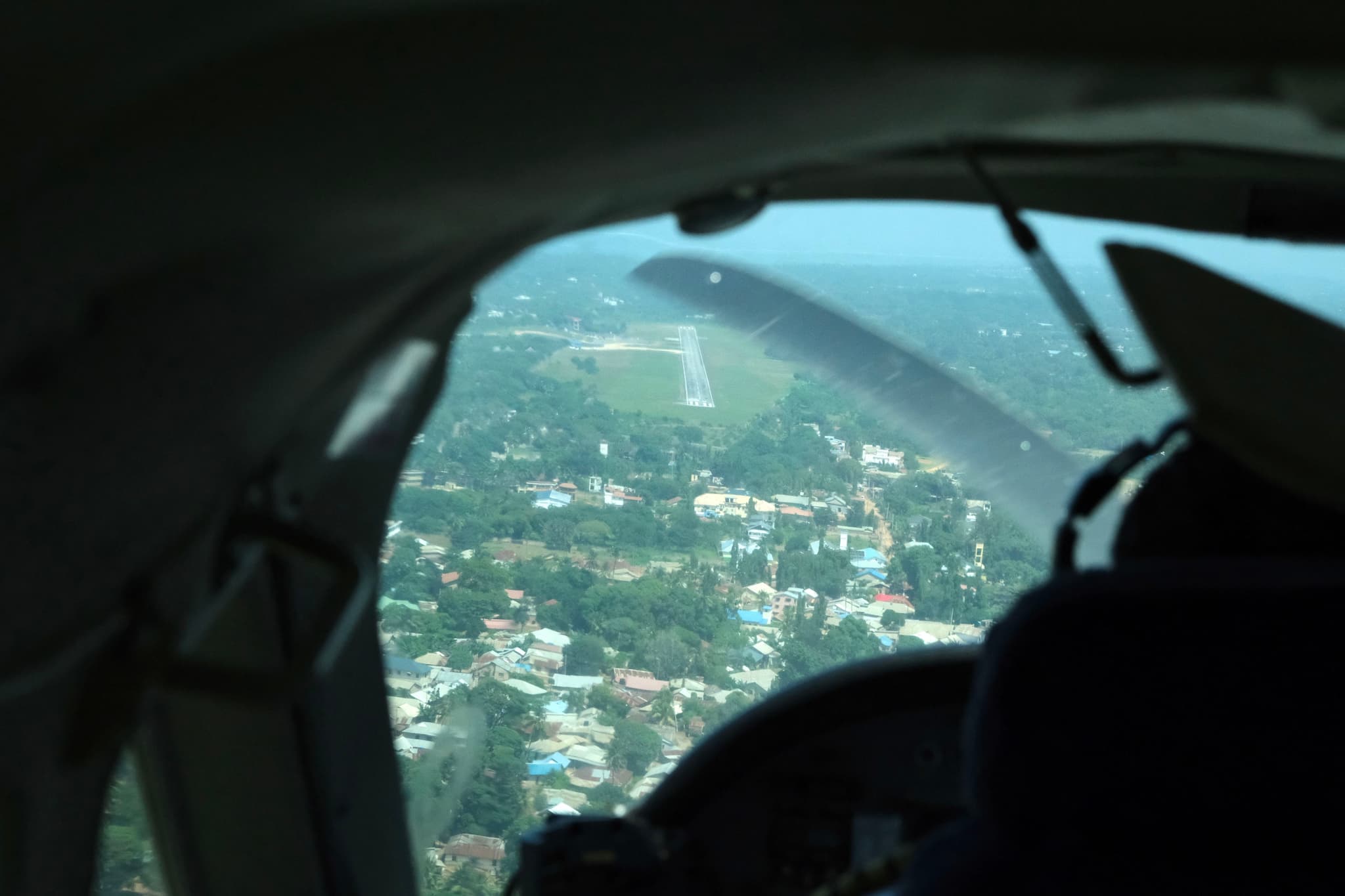 Aerial view from inside an aircraft cockpit, overlooking a landscape with a runway and surrounding greenery