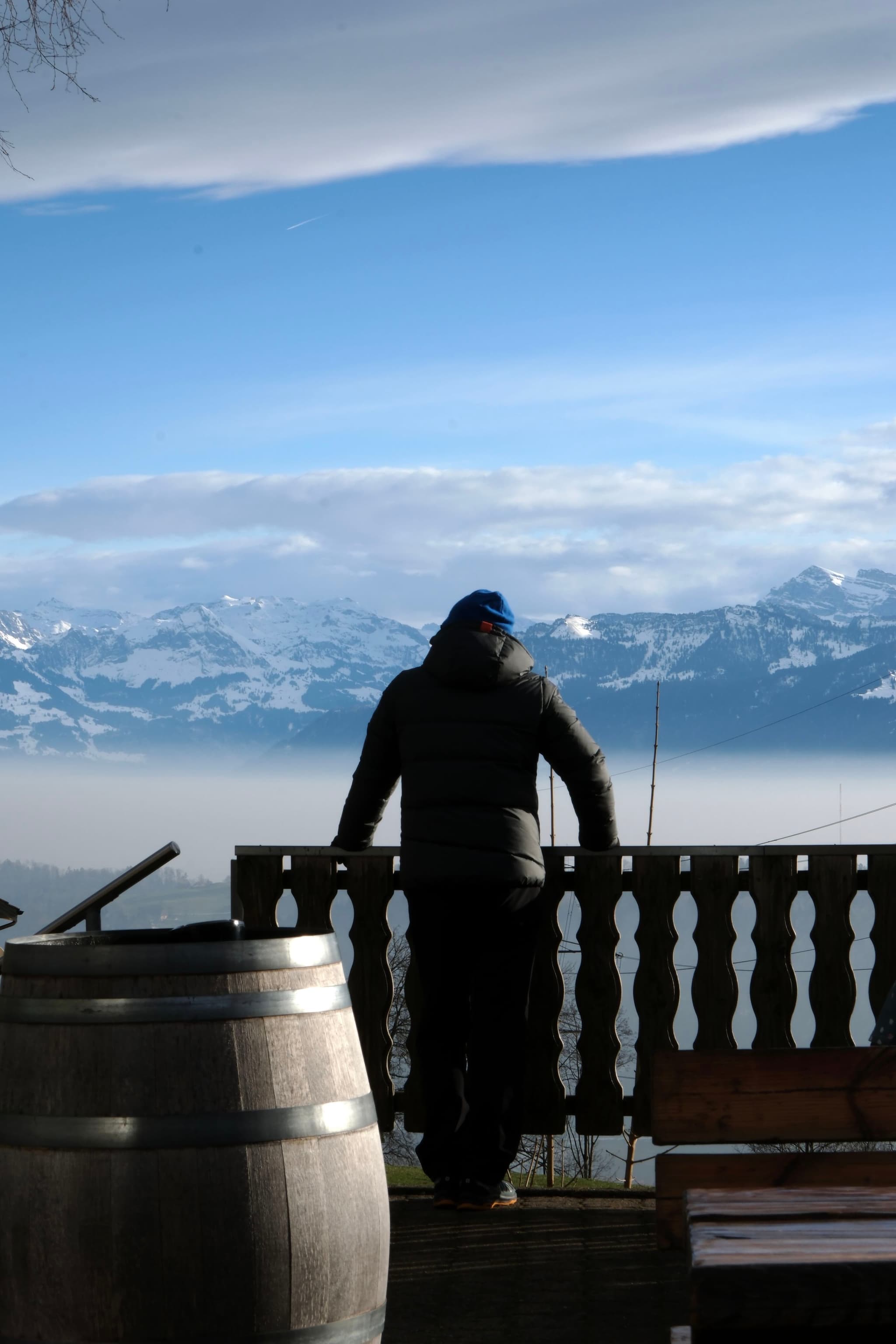 A person in a winter coat and hat stands on a balcony overlooking a snowy mountain range, with a wooden barrel nearby