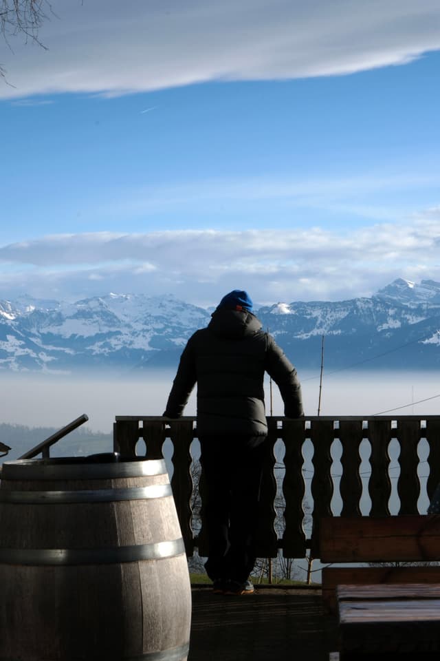 A person in a winter coat and hat stands on a balcony overlooking a snowy mountain range, with a wooden barrel nearby