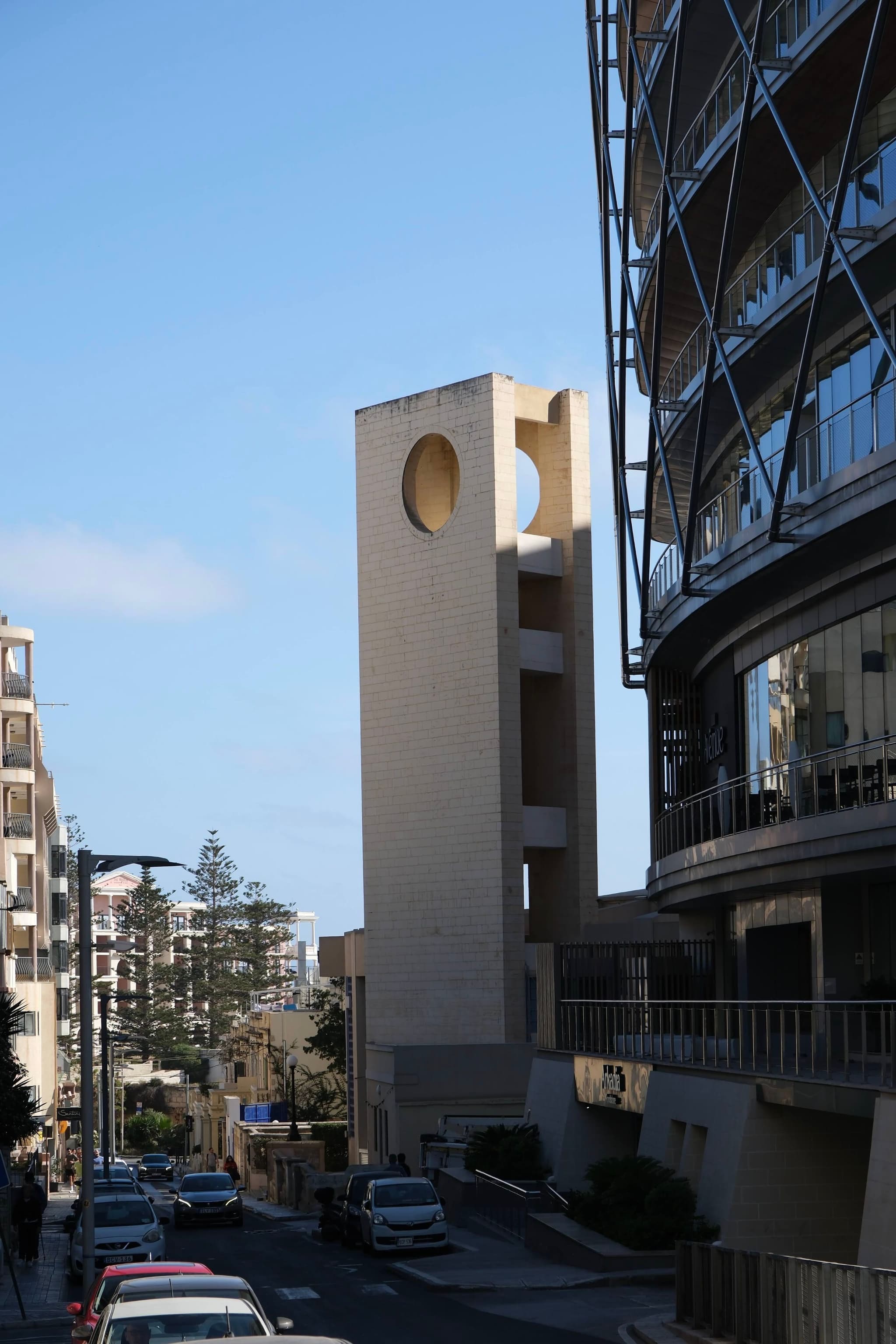 A street scene with modern buildings, including a tall structure featuring circular cutouts, under a clear blue sky