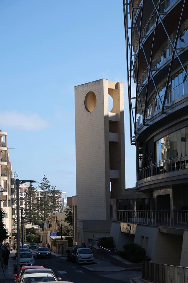 A street scene with modern buildings, including a tall structure featuring circular cutouts, under a clear blue sky