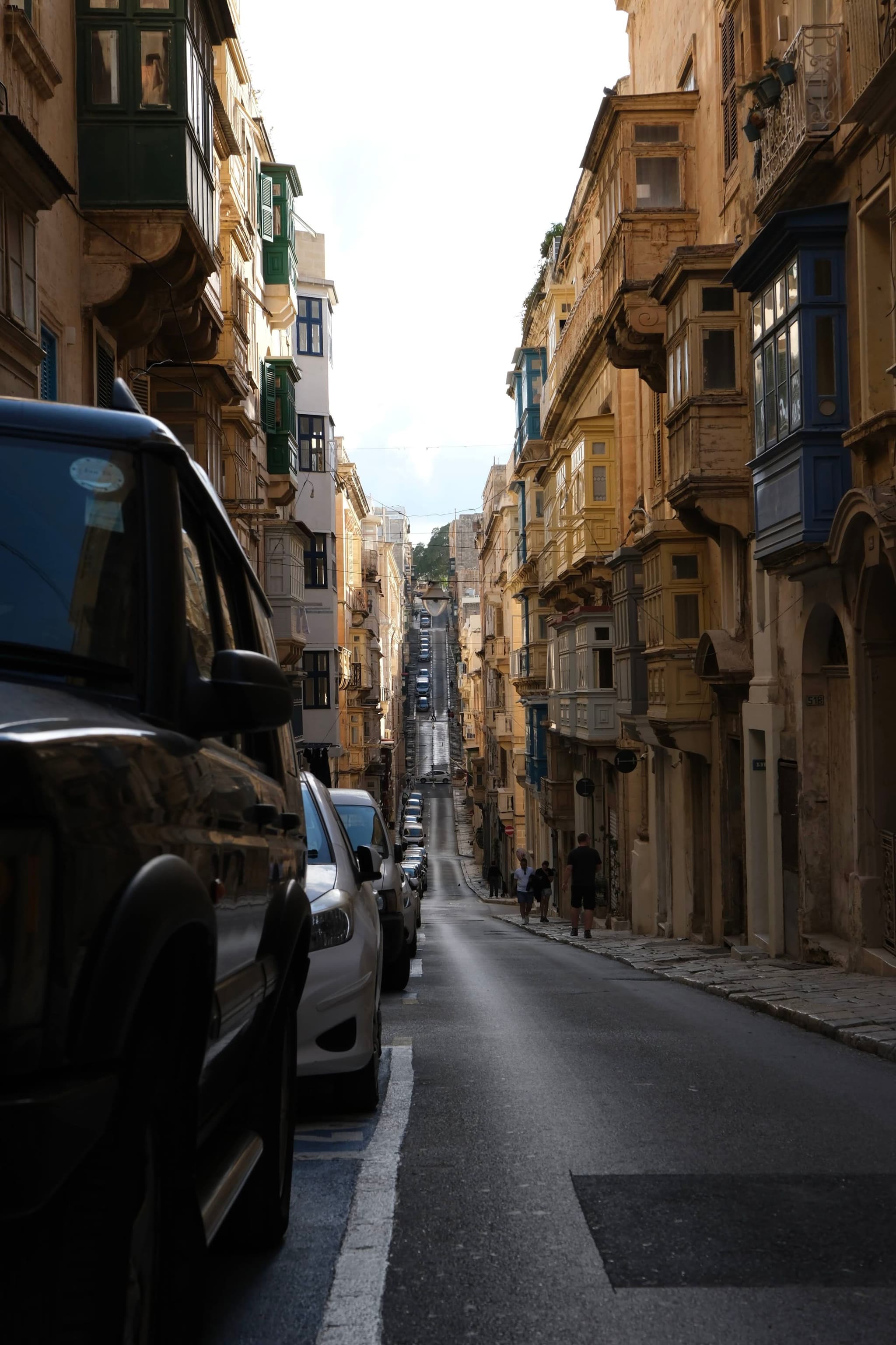 A narrow street lined with parked cars and historic buildings featuring traditional balconies, leading into the distance