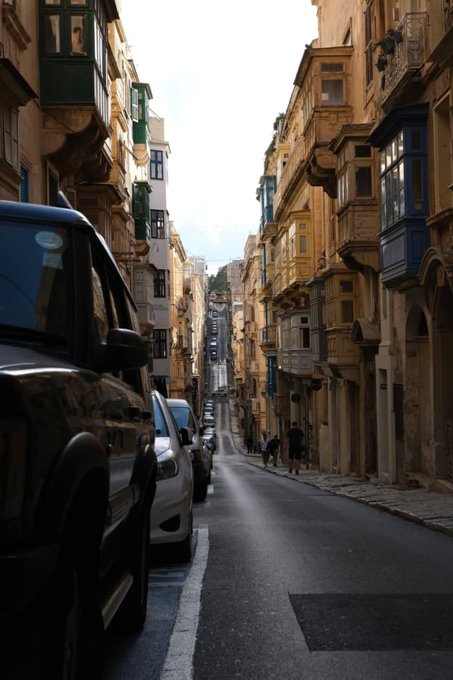 A narrow street lined with parked cars and historic buildings featuring traditional balconies, leading into the distance