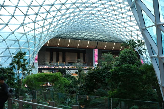 A large indoor space with a geometric glass ceiling, lush greenery, and a multi-level structure in the background