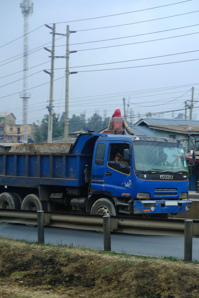 A blue Isuzu dump truck is driving on a road with a load of dirt. There are power lines and buildings in the background