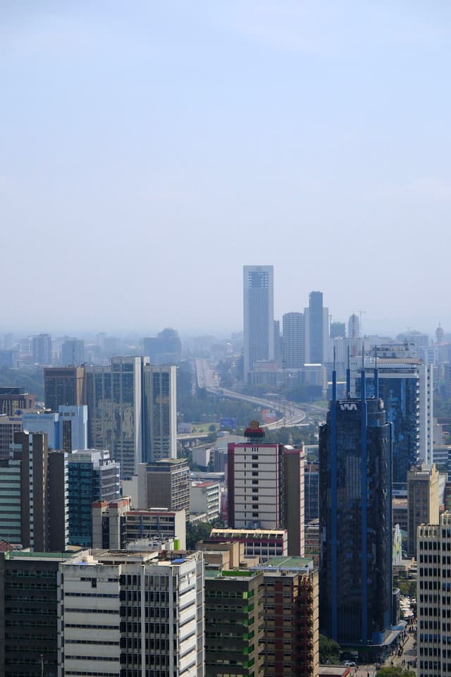 A cityscape featuring numerous skyscrapers and high-rise buildings under a clear blue sky, with a distant view of more buildings and a slightly hazy horizon