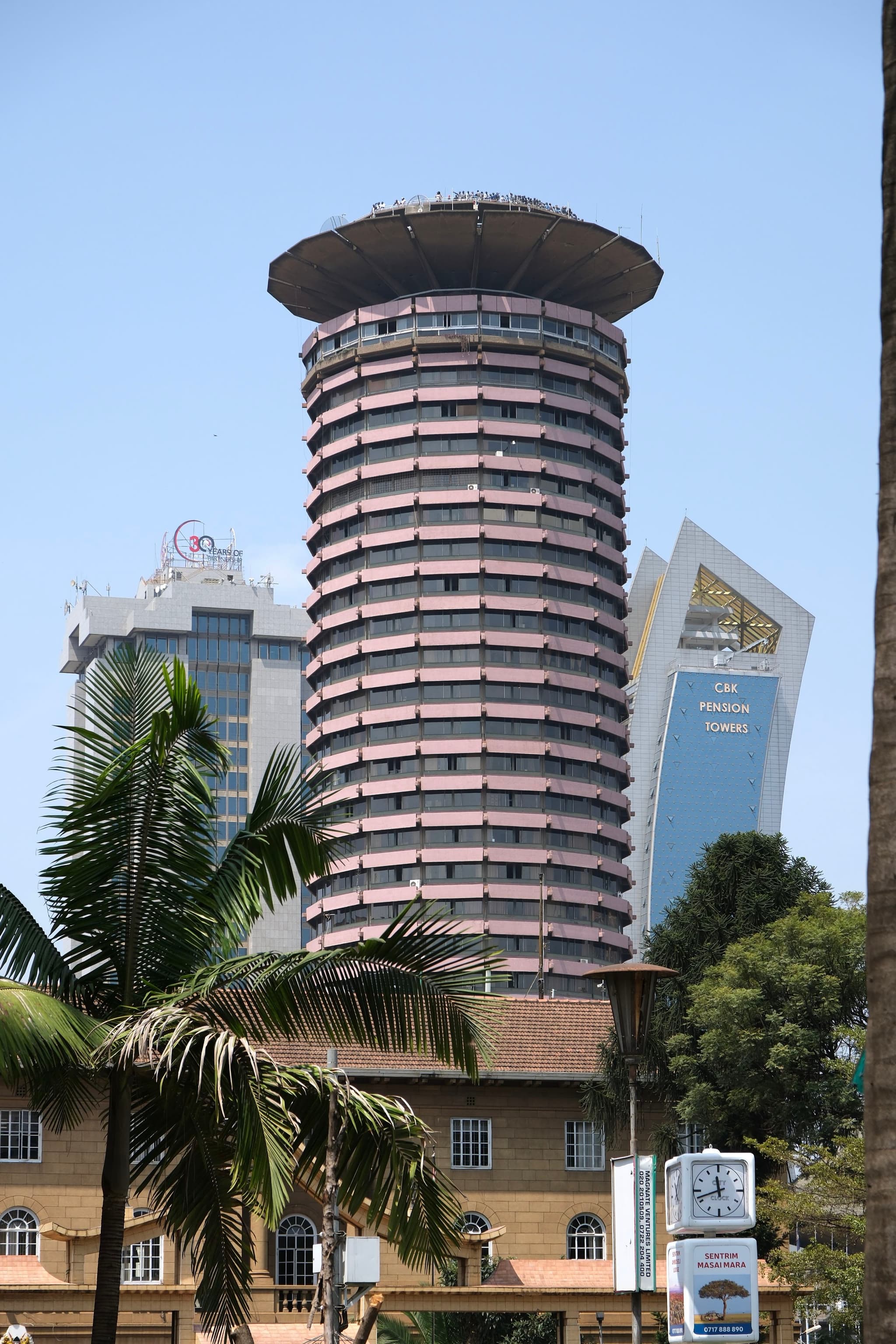 A tall, cylindrical building with a unique, tiered design stands prominently against a clear sky, surrounded by other modern structures and palm trees