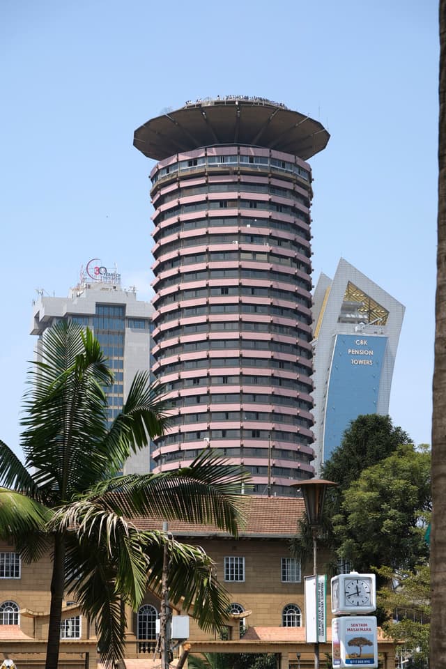 A tall, cylindrical building with a unique, tiered design stands prominently against a clear sky, surrounded by other modern structures and palm trees