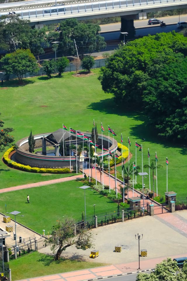 An aerial view of a landscaped park featuring a circular structure surrounded by flags on poles, with pathways and greenery