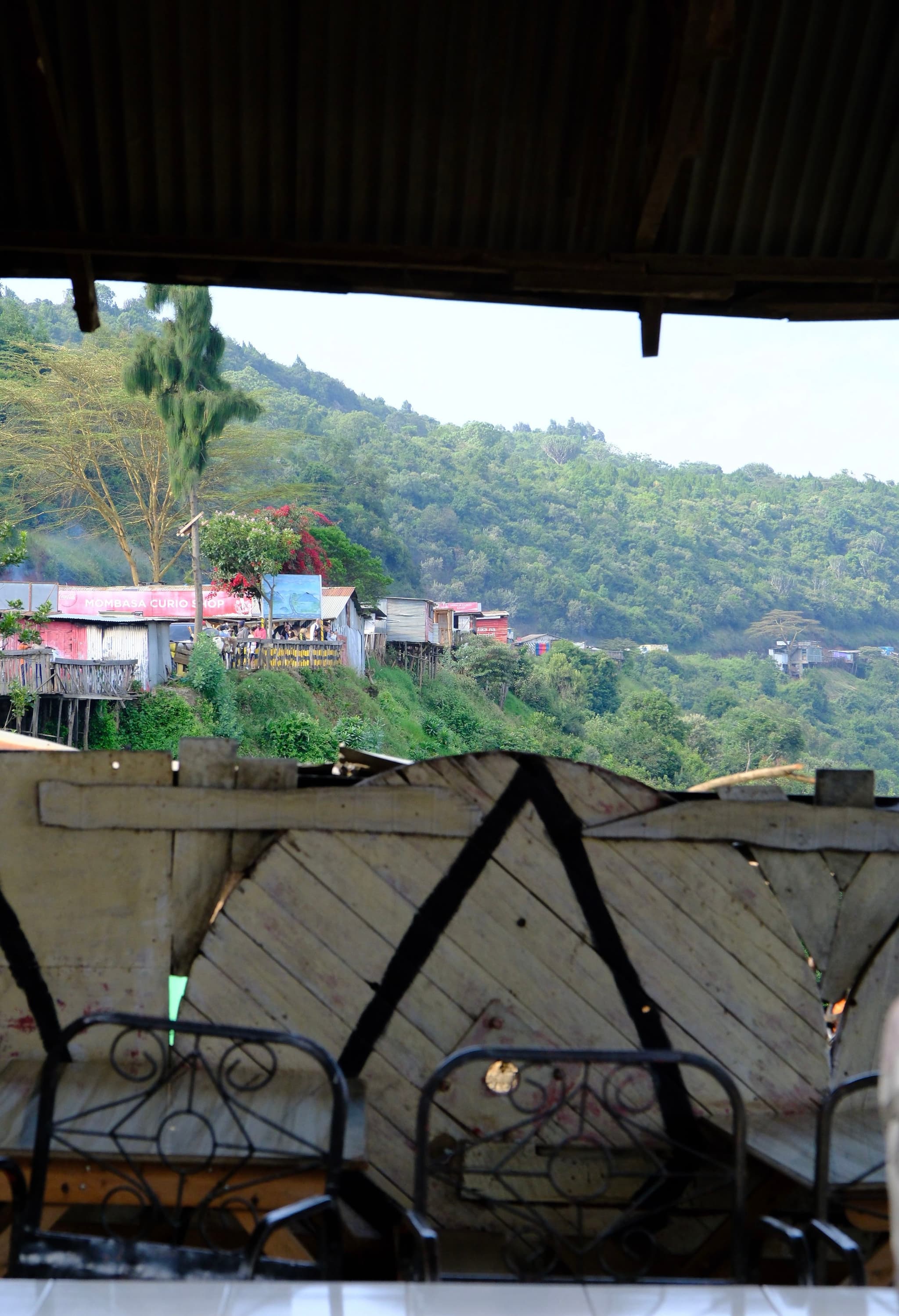 A view from a rustic, covered area with metal chairs, overlooking a lush, green hillside dotted with colorful houses