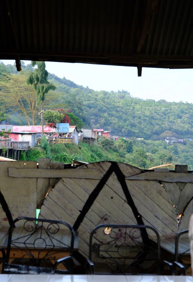 A view from a rustic, covered area with metal chairs, overlooking a lush, green hillside dotted with colorful houses