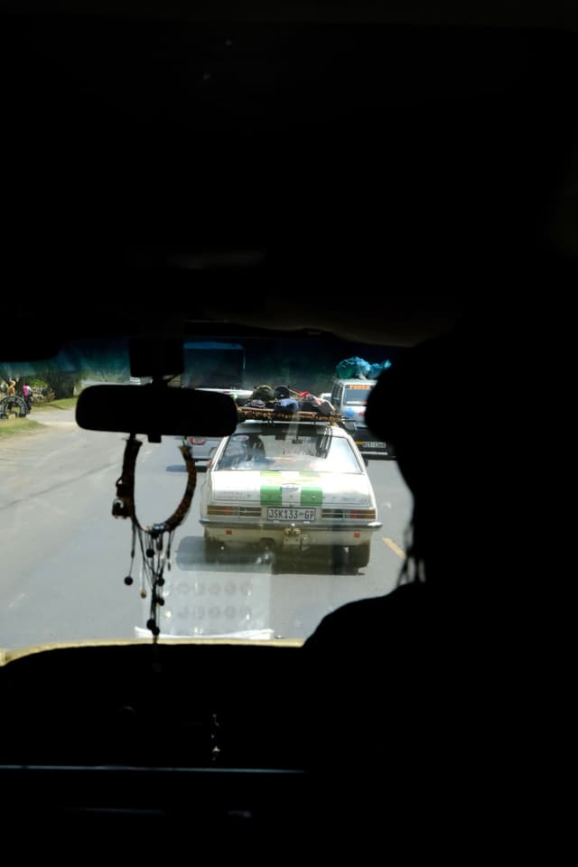 A view from inside a vehicle, looking out at a road with a car ahead. The silhouette of a person is visible on the right, and a rearview mirror with hanging decorations is in the foreground