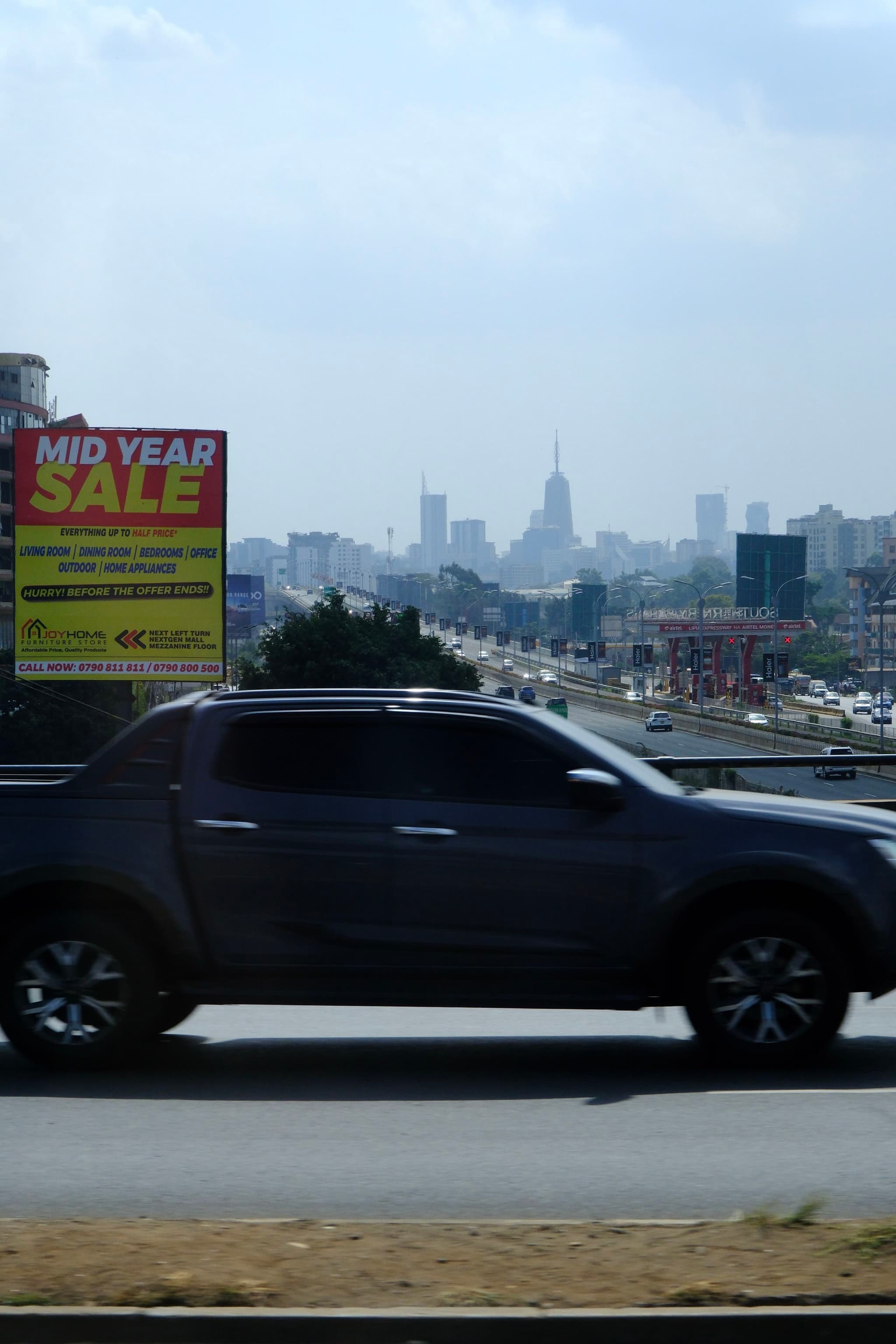 A dark-colored pickup truck drives on a highway with a city skyline in the background and a Mid Year Sale billboard on the side