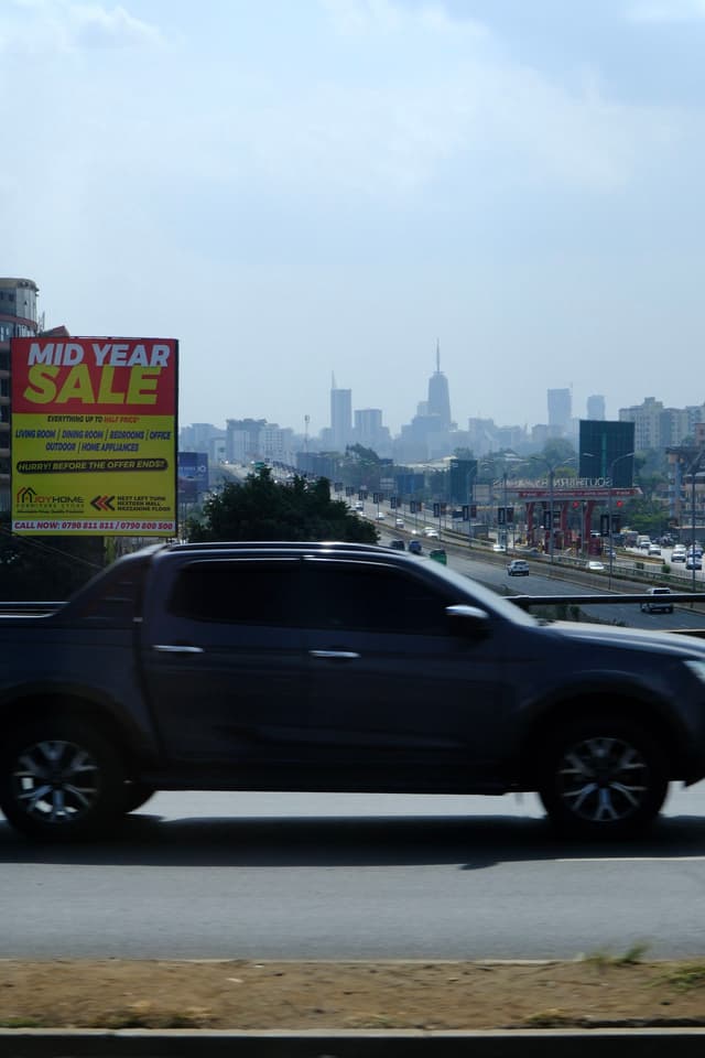 A dark-colored pickup truck drives on a highway with a city skyline in the background and a Mid Year Sale billboard on the side