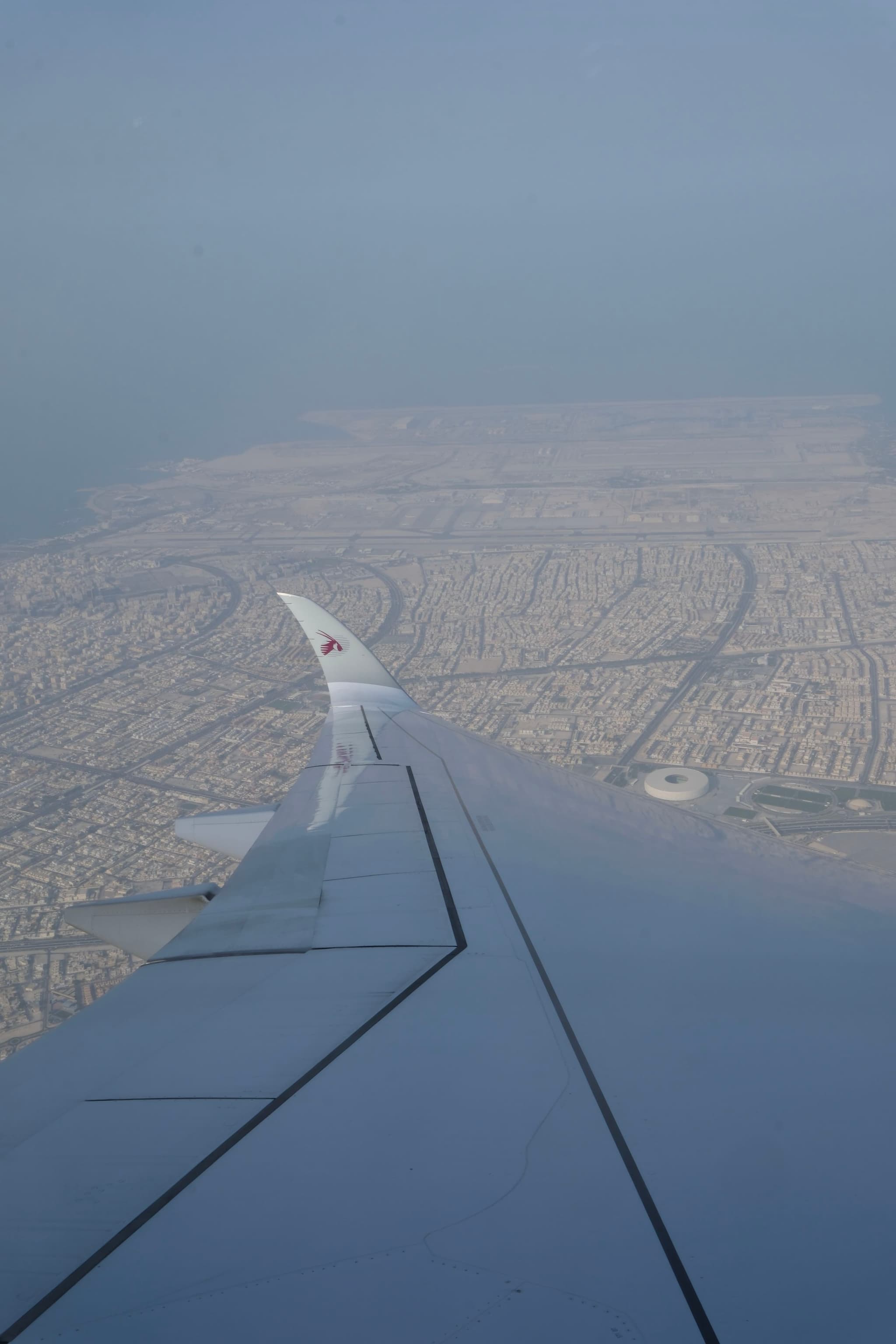 View from an airplane window showing the wing extending over a sprawling urban landscape with circular and grid-like patterns under a hazy sky