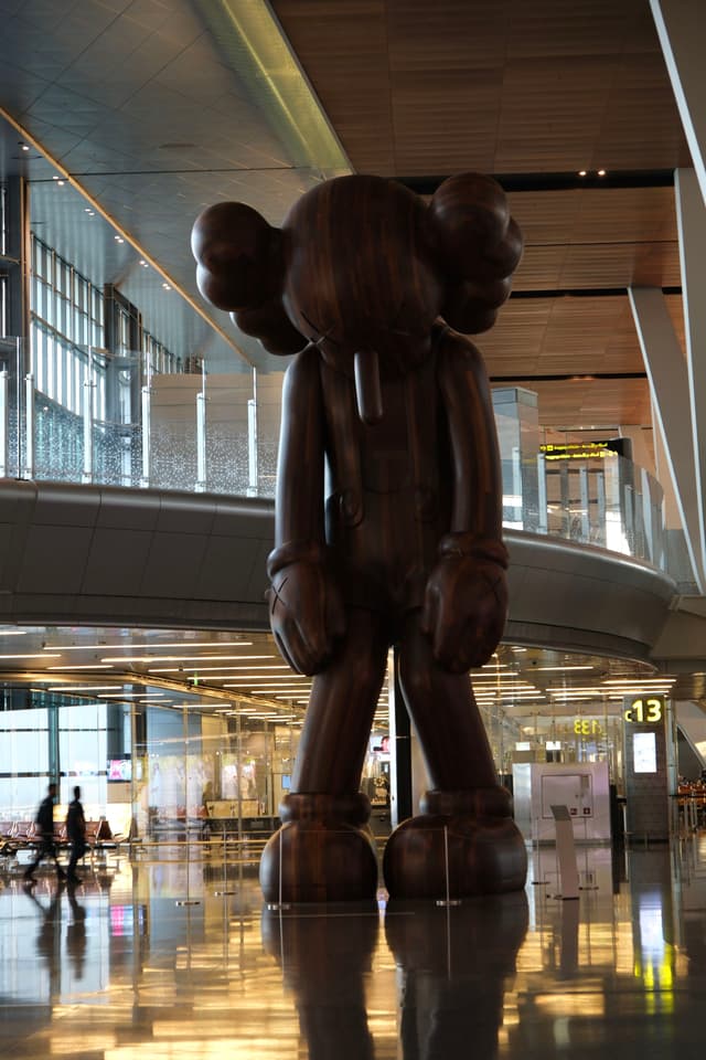 A large, dark sculpture of a cartoon-like figure with crossed-out eyes stands in an airport terminal, surrounded by modern architecture and reflective flooring