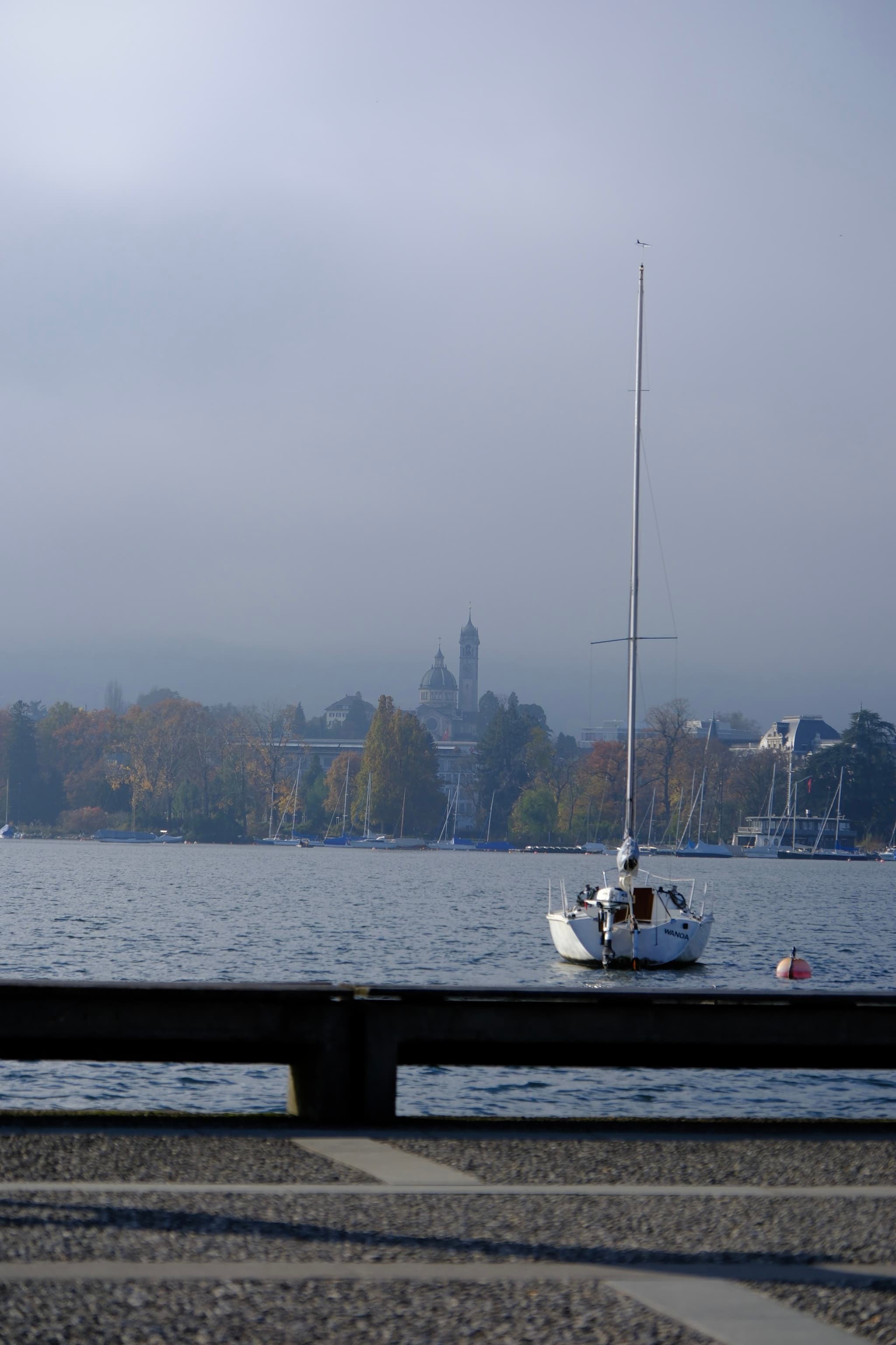 White sailboat moored on a calm lakeshore with distant tree line and faint city buildings under a hazy sky